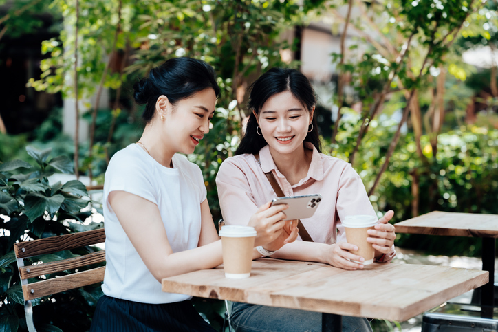 Two young Asian woman using smartphone and enjoying a coffee break against sunlight in garden. Having a coffee break. Work-life balance, mental wellbeing concept.