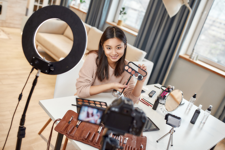 Asian female blogger showing cosmetic products while recording a tutorial video for her beauty blog using camera at home. Blogging, video blog, makeup and Work from Home concept